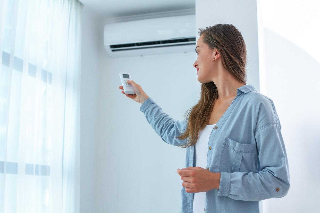 YOUNG WOMAN ADJUSTS THE TEMPERATURE OF THE AIR CONDITIONER USING THE REMOTE CONTROL IN ROOM AT HOME